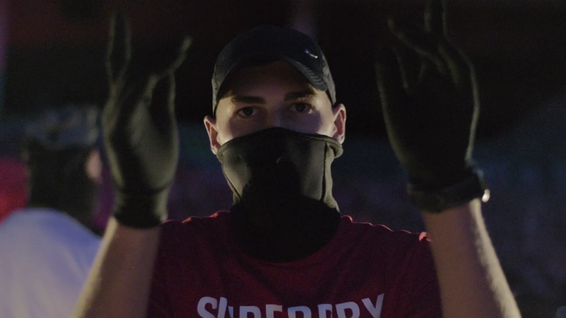 Man wearing a black cap, black face mask, black gloves, and a red shirt raising both hands.