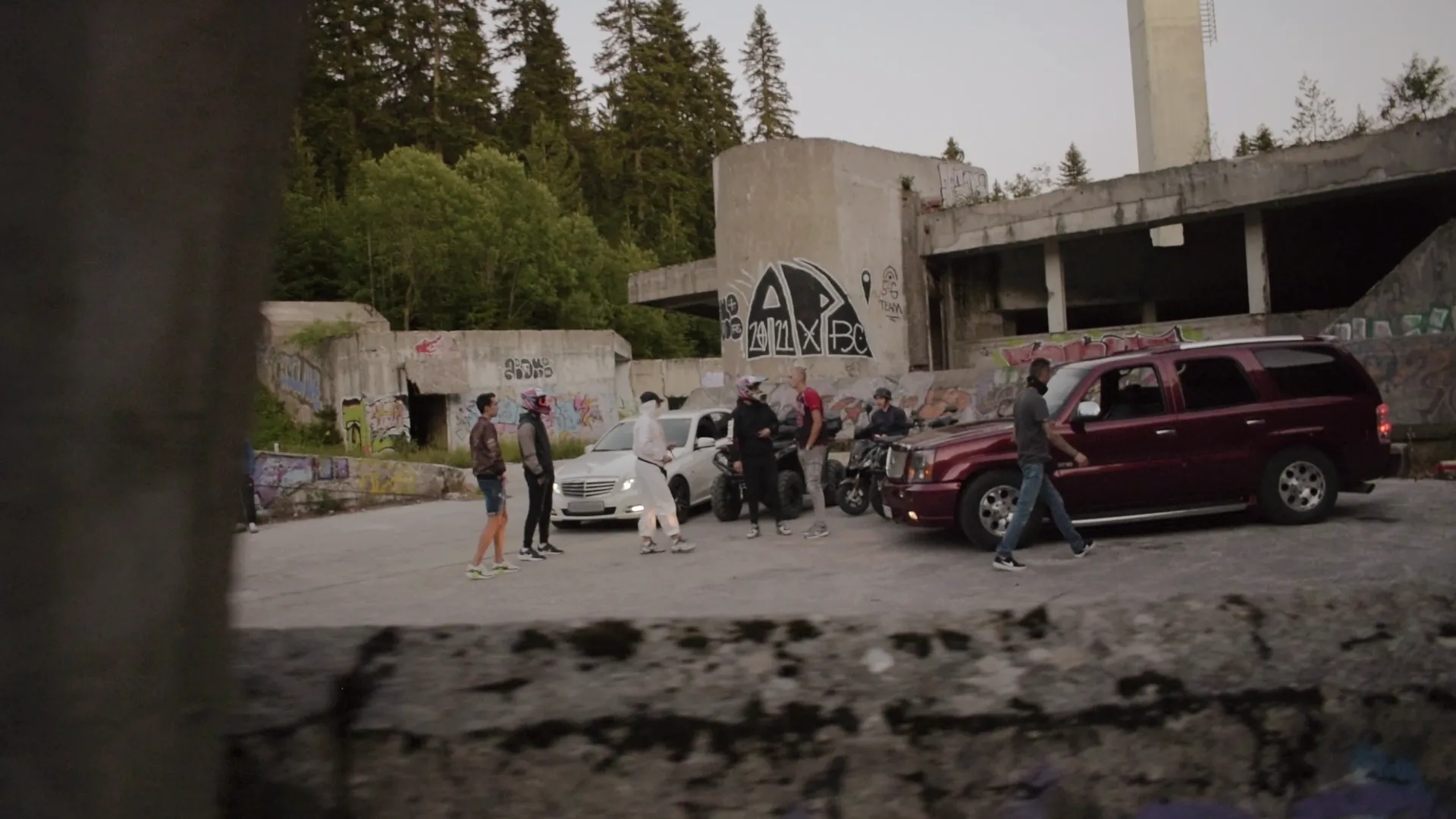 Group of people standing near a white car, a maroon SUV, and ATVs in an abandoned graffiti-covered concrete area surrounded by trees.