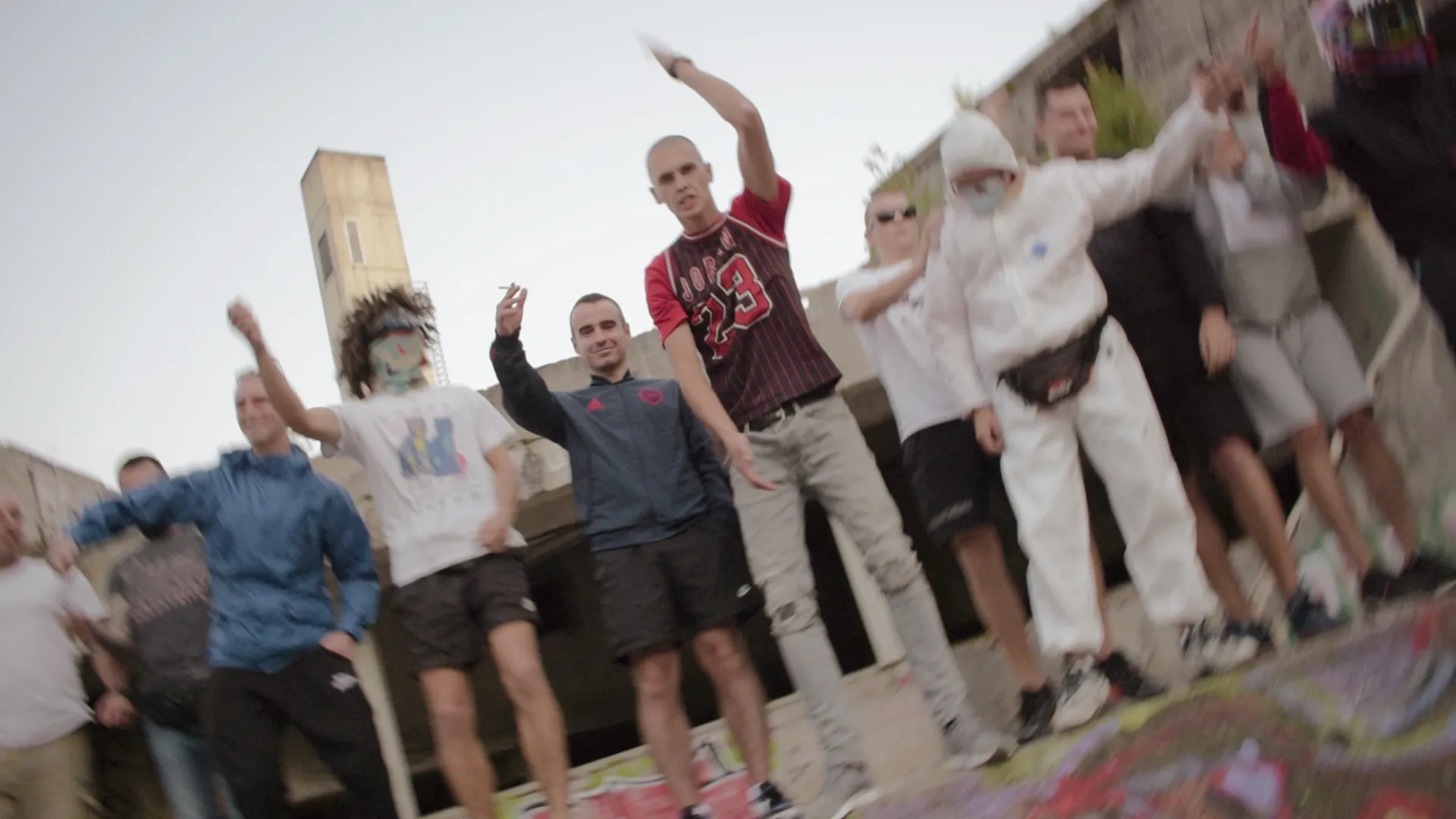 Group of young men standing outdoors near concrete structures, some raising their arms and one wearing a white hooded outfit and mask.
