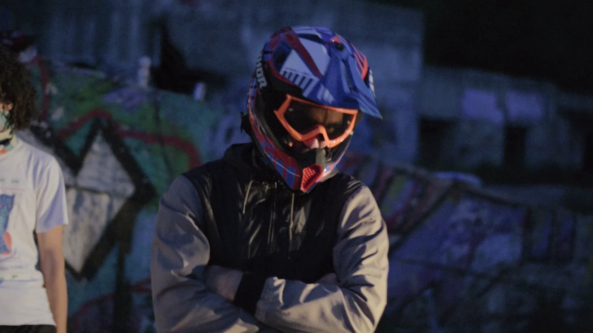 Person wearing blue and red motocross helmet with orange goggles and a jacket, standing with arms crossed in front of graffiti-covered walls at night.