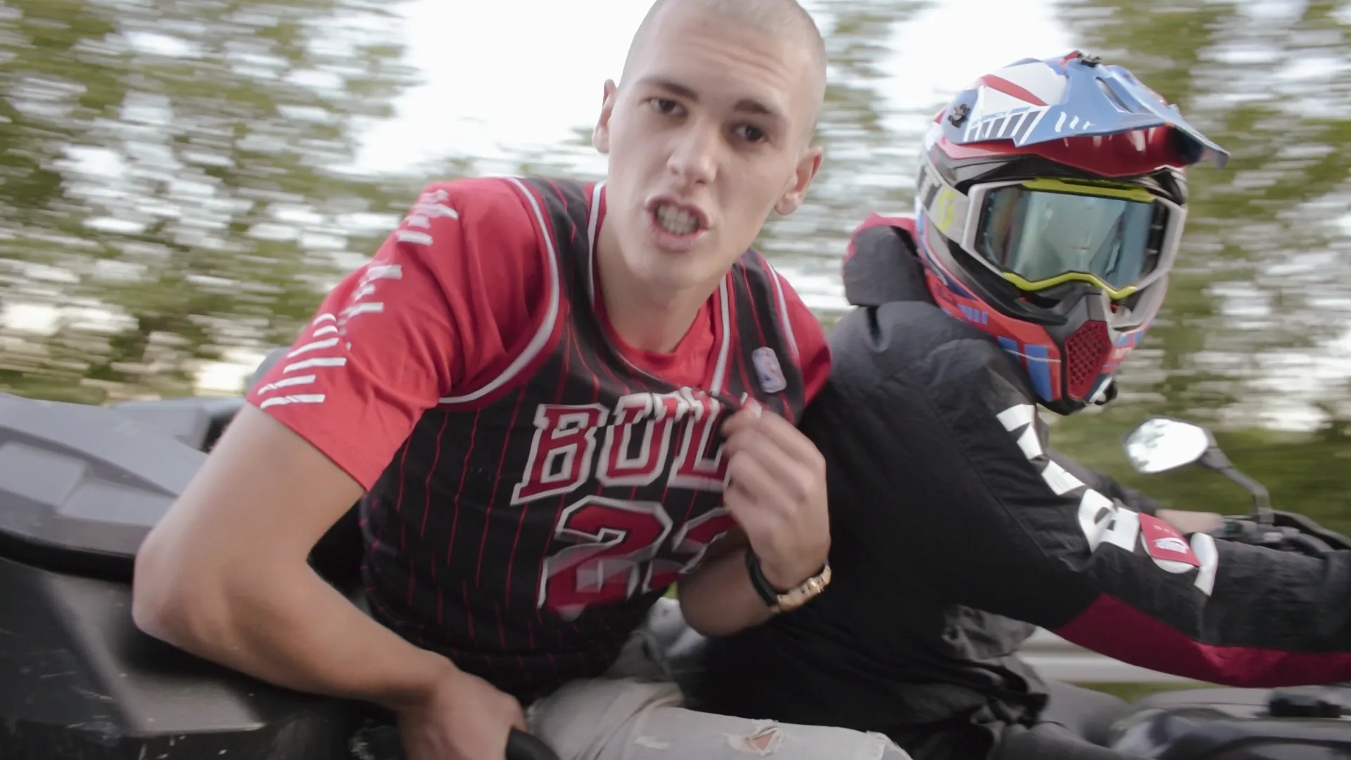 Two young men riding a motorcycle, one wearing a red Chicago Bulls jersey and the other wearing a colorful helmet and black jacket.