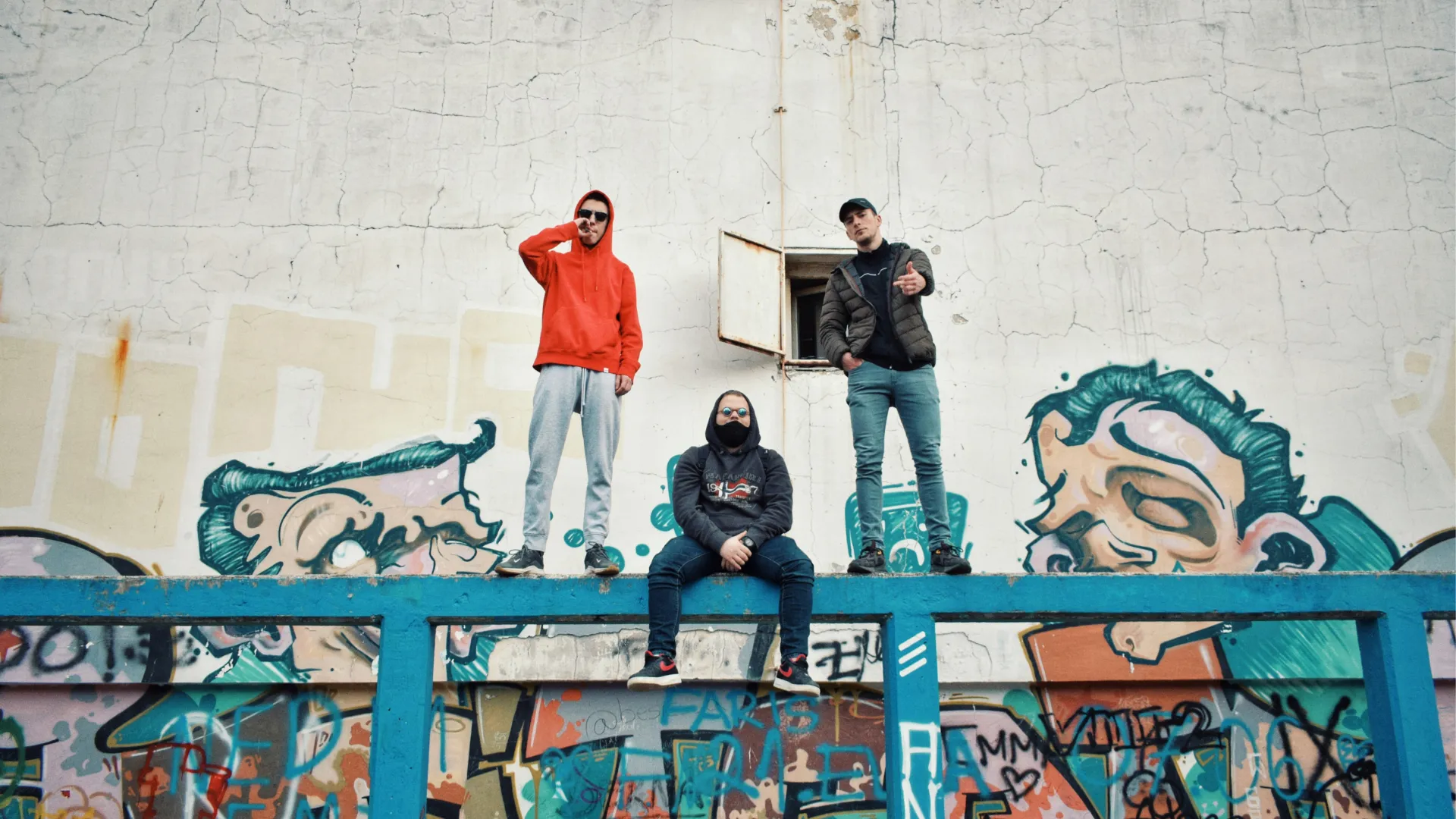 Three young men posing on a blue metal railing in front of a graffiti-covered wall featuring cartoonish faces.