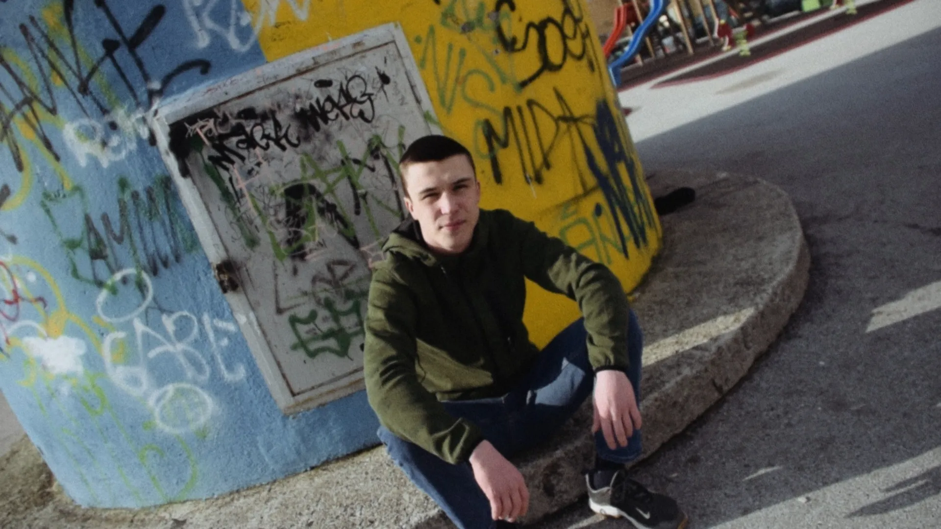 Young man in green jacket and blue jeans sitting on a concrete ledge in front of graffiti-covered blue and yellow walls.