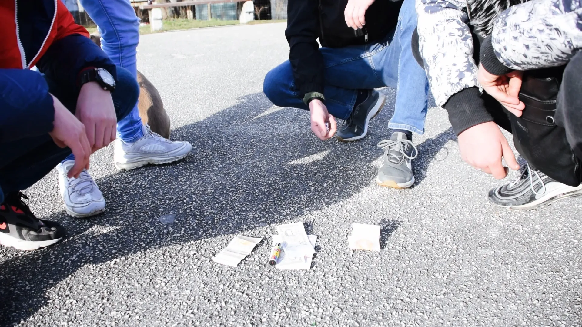 Four people crouching outdoors around scattered currency notes and a small colorful object on a gravel surface.
