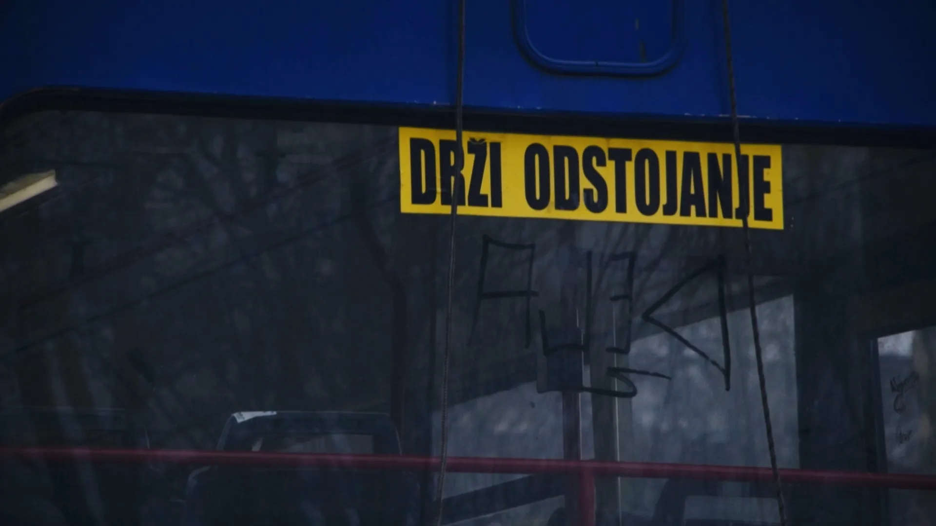 Yellow sign with black text 'DRŽI ODSTOJANJE' on a dark tinted bus window with reflections of trees.