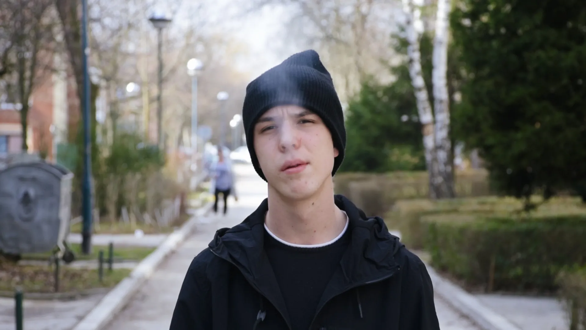 Teenage boy wearing a black beanie and jacket standing on a tree-lined sidewalk with smoke near his face.