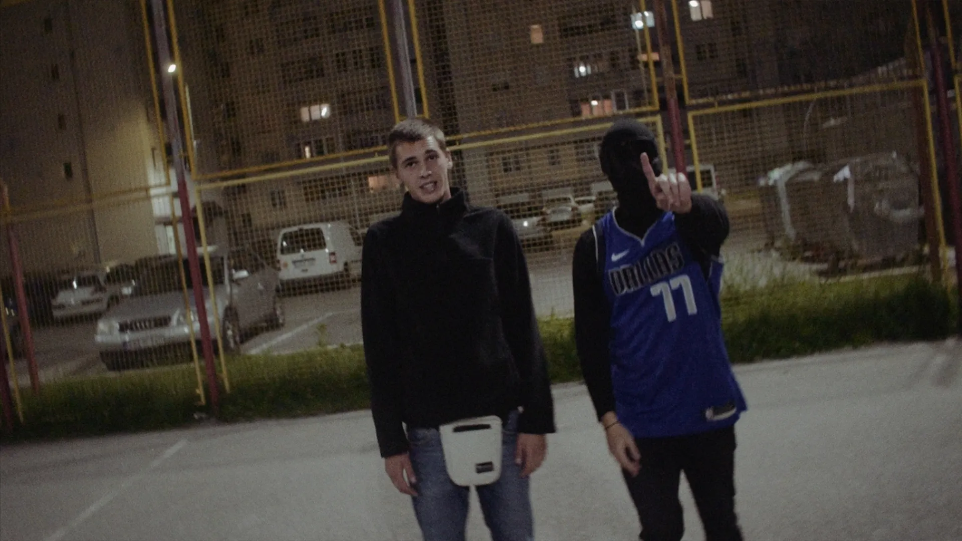 Two young men standing outside at night near a fenced parking lot, one wearing a blue basketball jersey and a black face mask raising his hand, and the other in a black jacket with a white pouch.