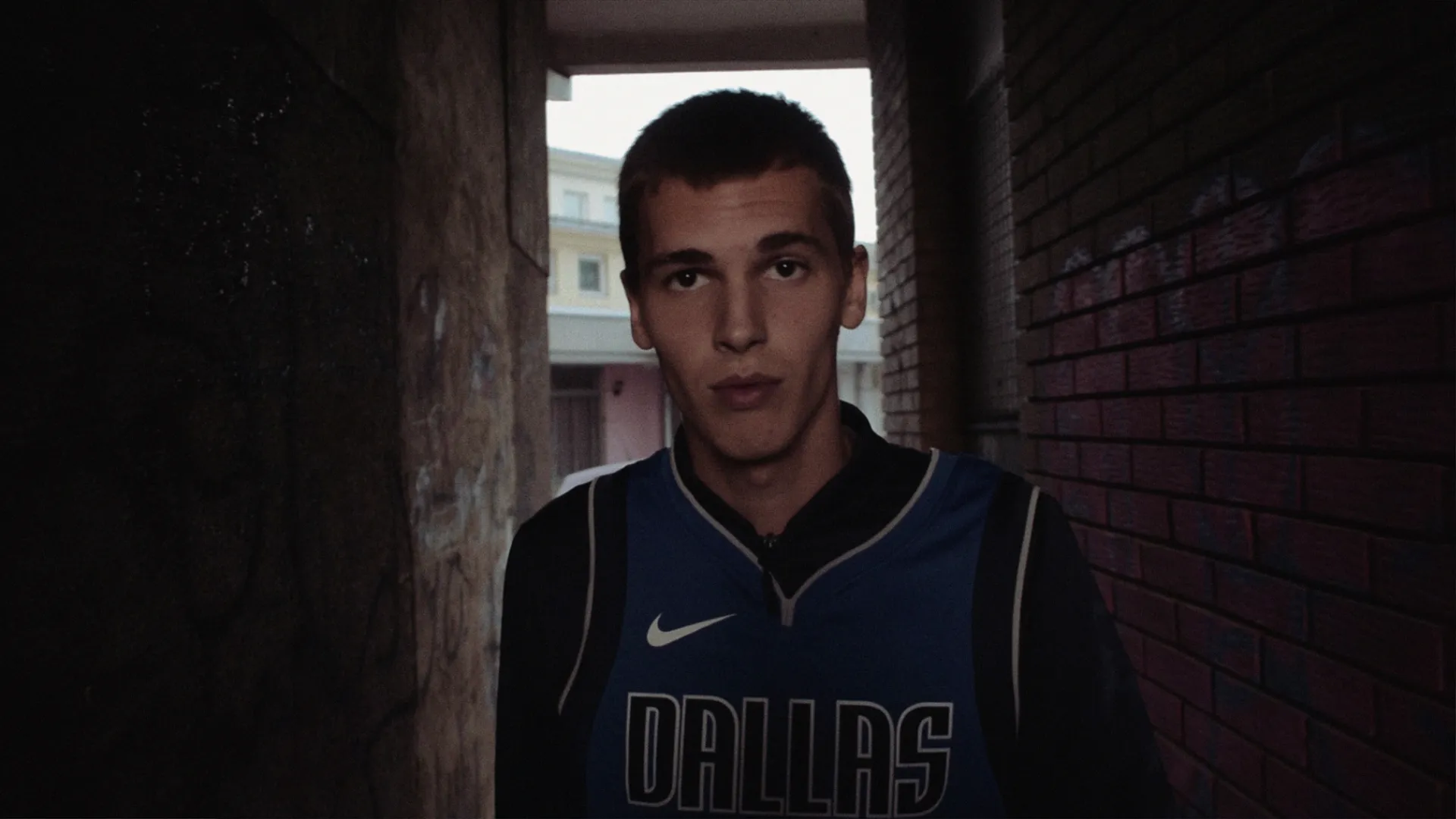 Young man wearing a Dallas basketball jersey standing in a narrow alleyway between a graffiti-covered wall and a brick wall.