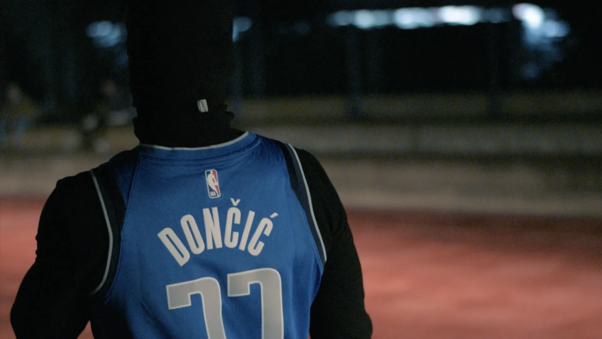 Person wearing a blue basketball jersey with the name Dončić and number 77 on the back, standing on a dimly lit court at night.